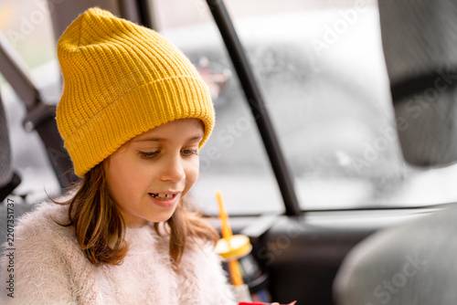 Portrait of a smiling little girl wearing a yellow knitted beanie while sitting inside a car during a family trip or everyday travel.