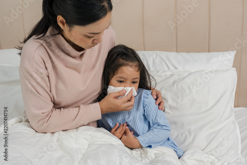 mother helping sick preschoool child girl blowing nose with tissue paper in bed