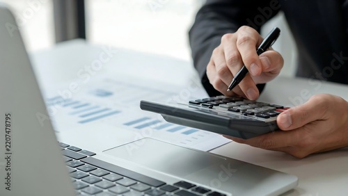 Businessman using a calculator and reviewing financial data at an office desk with a laptop