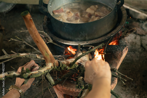 Woman hands feeding dry firewood branches into a traditional clay stove fire under a simmering food pot
