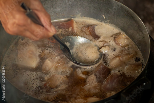 Hand using a metal ladle to skim foam from a boiling pot of simmering pork meat broth for clear soup