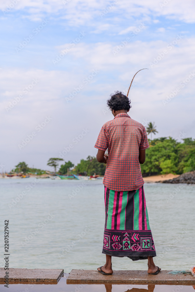 Fototapeta premium A traditional fisherman stands on a coastal pier holding a long fishing rod, facing calm tropical waters and a lush shoreline