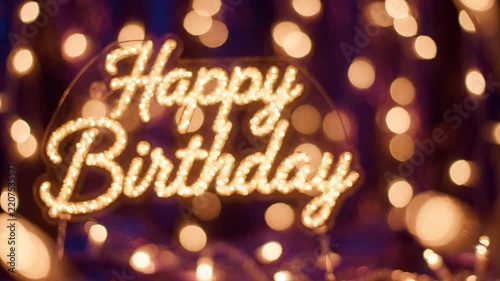 A glowing 'Happy Birthday' sign illuminated with warm lights against a blurred background of bokeh lights, creating a festive and celebratory atmosphere.
