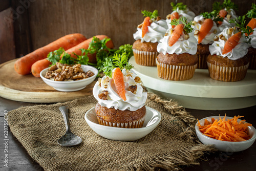 Freshly baked carrot cake cupcakes topped with smooth buttercream, walnuts, and fresh parsley, served on a rustic burlap tablecloth alongside raw carrots