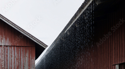 Rainwater falling continuously from the wooden eaves of two adjacent red barns forming a solid vertical curtain during a storm