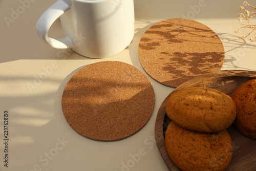 Cork round coasters and oatmeal cookies, ceramic cup on beige background with shadow from sunlight