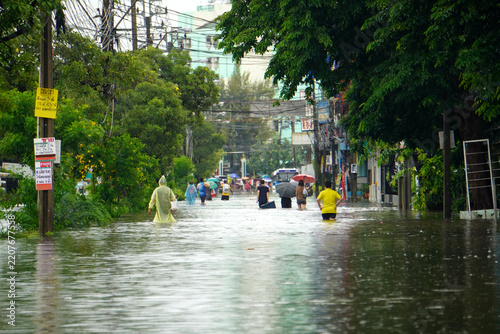 Fotografie Bangkok, THAILAND - September 27, 2026 : Amidst the rising floodwaters on the streets, people are walking out using umbrellas and wearing raincoats to buy food
