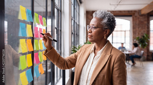 Senior African American businesswoman writing on colorful sticky notes on glass wall while planning strategy