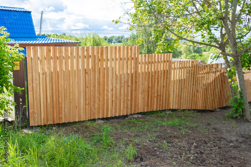 peaceful wooden fence installed in serene outdoor residential area with blue-roofed house and green tree in background. side view of natural setting with clean concrete and brown dirt.