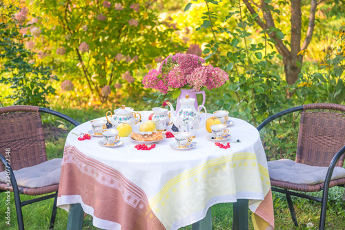 Summer breakfast in the blooming garden; coffee cups, coffee pot, berries, apples and bouquet of pink hydrangea flowers on the table covered by linen tablecloth in the sunny summer garden