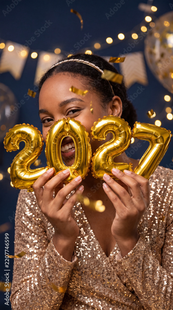 Fototapeta premium Woman holding golden 2027 number balloons while wearing a sparkling sequin top at a festive indoor celebration