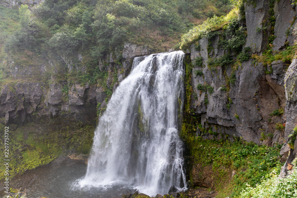 Obraz premium Scenic view of Snow Leopard waterfall flowing over a rugged rock cliff into a calm pool, surrounded by lush green forest vegetation and mossy stone walls