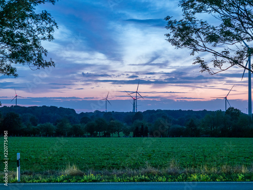 Wind turbines silhouetted against a colorful dusk sky over green fields and forest at twilight
