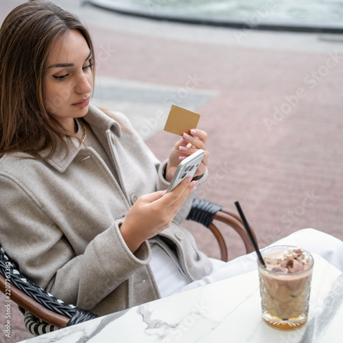 Young Gen Z business woman holding gold credit card and using smartphone at outdoor cafe. Female paying online, shopping or managing mobile banking account