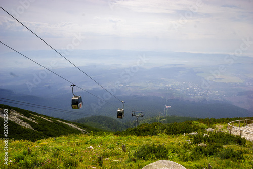Cable cars ascend a mountain slope with a village in the valley below on a hazy day.