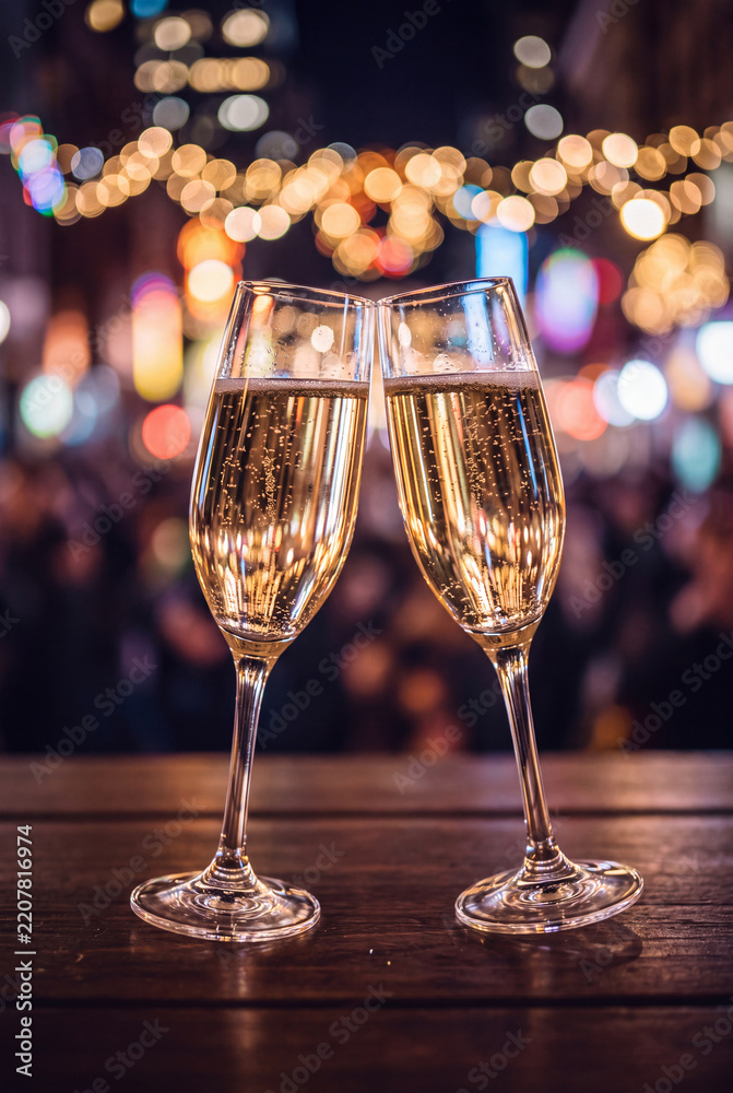 Obraz premium Two elegant glasses of sparkling champagne placed on an outdoor wooden table with colorful glowing urban bokeh lights shimmering in the background