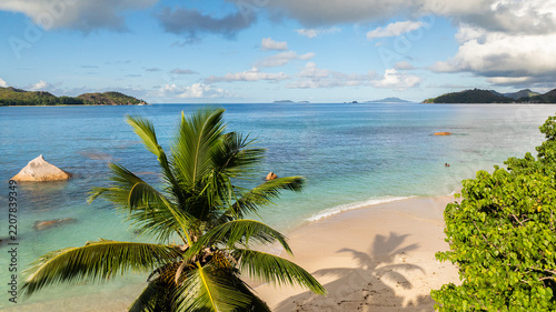 Beach lined with palm trees, calm turquoise waters, and distant green islands on the horizon. Praslin, Seychelles.