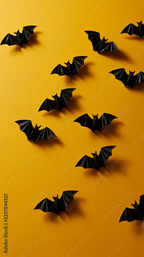 Fototapeta premium Flat lay of black folded paper bats arranged in an expanding arc across a cheerful golden yellow background sharply creased wings casting delicate soft shadows and leaving