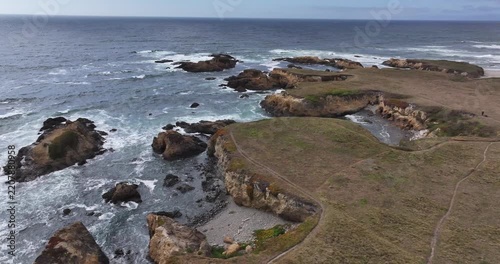 Aerial view of the rugged coastline and rocky cliffs at Glass Beach with crashing waves and grassy trails in Fort Bragg, California, United States.