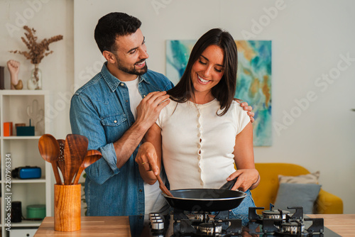 A warm scene shows a couple enjoying food prep at a stove, one stirs a skillet while the other smiles and assists. Wooden utensils stand in a holder, emphasizing teamwork and shared cooking