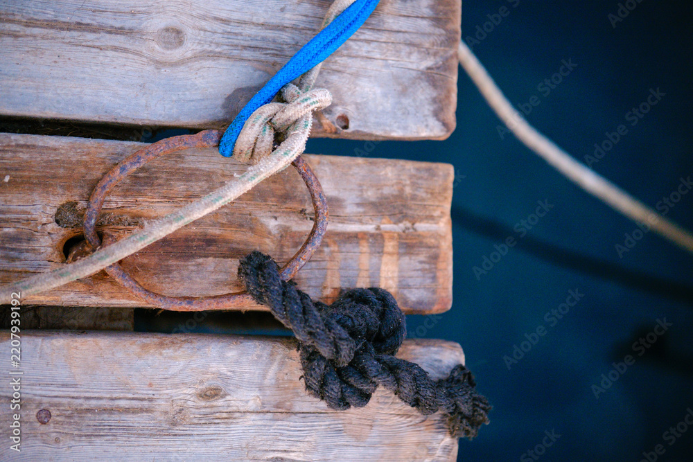 Fototapeta premium Rusty metal mooring ring on aged wooden pier boards with tied ropes and nautical knots over calm blue ocean water surface