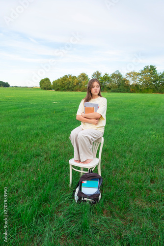 A simple white wooden chair and a black backpack with books standing on lush green grass in a peaceful garden. Back to school concept.