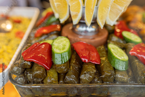 Close up view of traditional Turkish sarma or dolma stuffed grape leaves garnished with fresh cucumber slices, red bell pepper, and lemon wedges at buffet