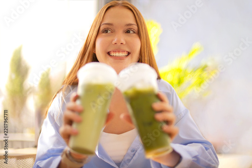 Smiling woman holding two green smoothies toward camera with bright fruit decor and softly blurred indoor background, fresh beverage portrait in a sunlit interior setting