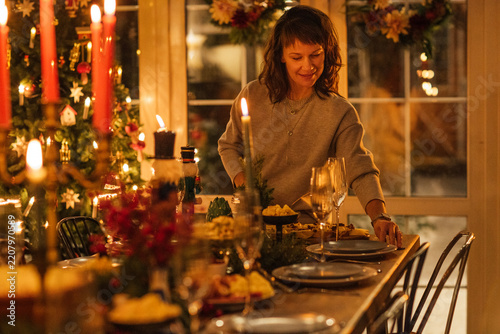 Woman setting a festive dining table with plates and decorations for a holiday meal, Christmas tree and candles visible in a cozy indoor setting