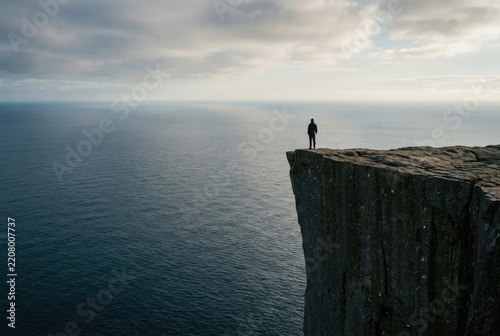 A lone figure stands on the edge of a towering cliff overlooking a vast, calm ocean under a cloudy sky.