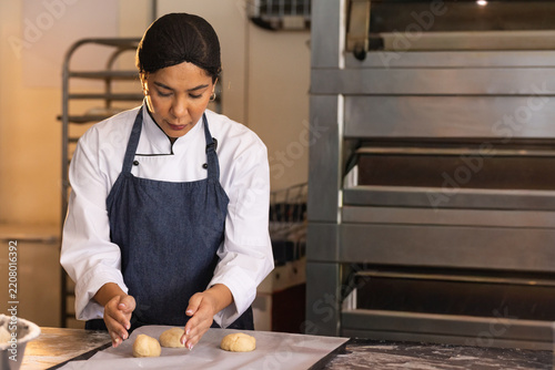 Female baker in chef jacket and navy apron shaping three dough portions on bakery prep table
