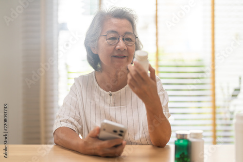 Asian senior woman reading medicine label on a bottle holding smartphone at home, Elderly female checking vitamins and supplements for medication management, Healthy aging digital healthcare concepts