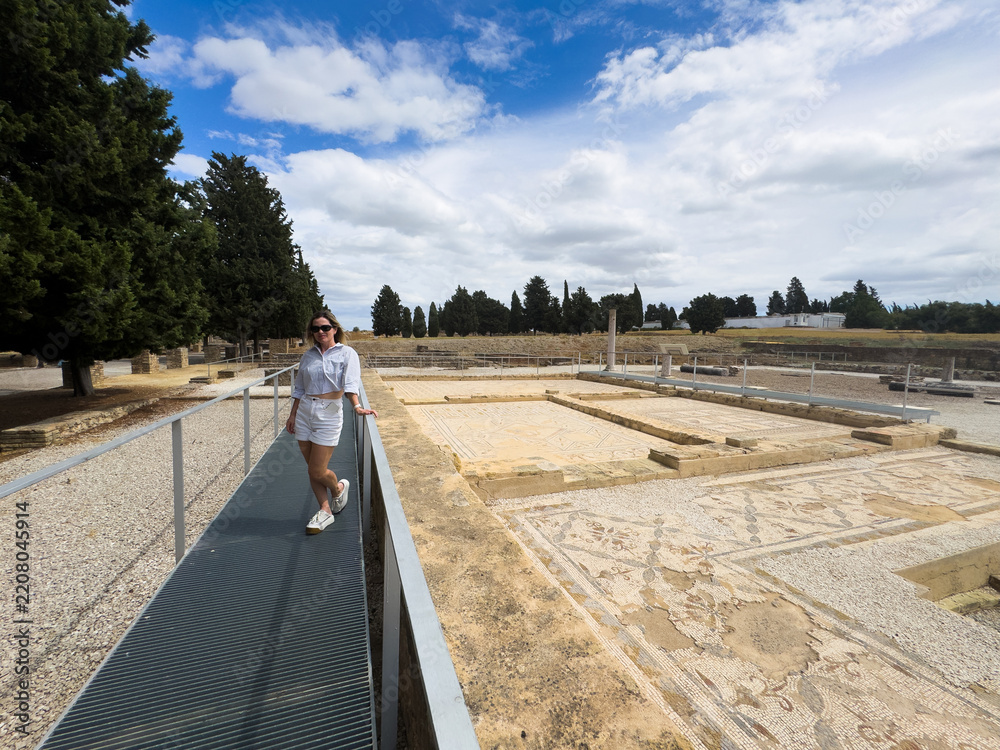 Obraz premium woman exploring ancient ruins with mosaic floors and stone columns, wide angle shot from a slight elevation, blue sky and white clouds in the background, modern and serene atmosphere, ideal.