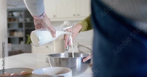 Tilting carafe is pouring milk while whisking hand is mixing bowl to make batter on countertop