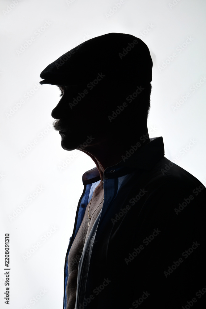 Naklejka premium Dramatisches Silhouettenporträt eines älteren Mannes mit Schiebermütze und Sakko im Seitenprofil. Low-Key, Schatten, Charakter, Vintage-Stil, geheimnisvolle Stimmung und Studiofotografie.