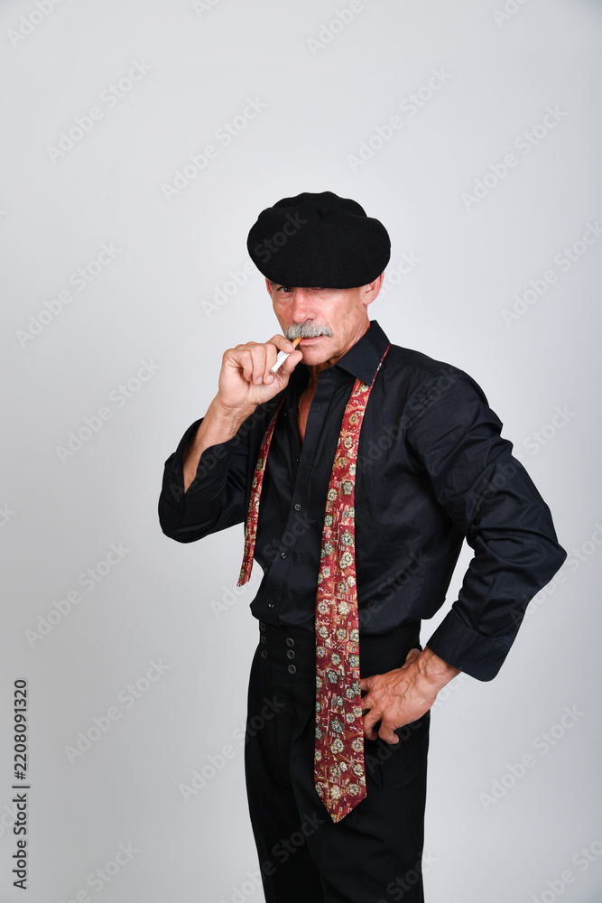 Naklejka premium Charakterporträt eines älteren Mannes mit schwarzer Schiebermütze, Hemd und gemusterter Krawatte, der nachdenklich die Hand ans Gesicht hält. Vintage-Stil, Persönlichkeit und Studiofotografie.