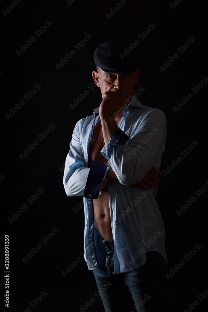 Naklejka premium Dramatisches Low-Key-Porträt eines Mannes mit schwarzem Hut und offenem Hemd vor dunklem Hintergrund. Nachdenkliche Pose, Schatten, Charakter, Mystery, Vintage-Stil und künstlerische Studiofotografie.