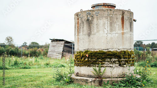 Old rustic concrete water tank standing in overgrown grassy field near abandoned wooden shed