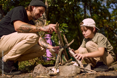 Smiling dad and boy build campfire together near lake at forest campsite. Family bonding, outdoor cooking and camping tradition concept.
