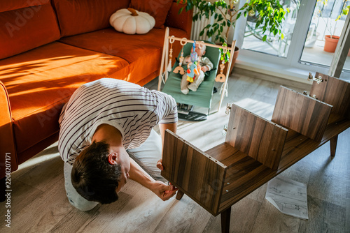 Father building new tv stand furniture on the floor, carefully following instructions while his baby watches from a bouncer in the brightly lit living room, creating a family home