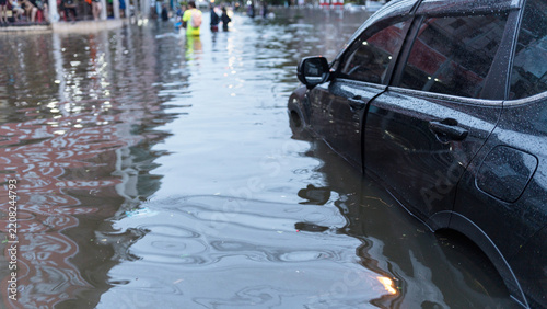A dark car parked on a flooded city street during a heavy rainstorm with people in the distance