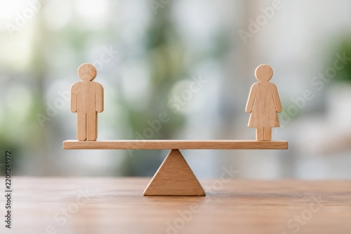 Wooden figures of a man and a woman balanced equally on a seesaw, symbolizing gender equality and balance in a neutral, blurred background.