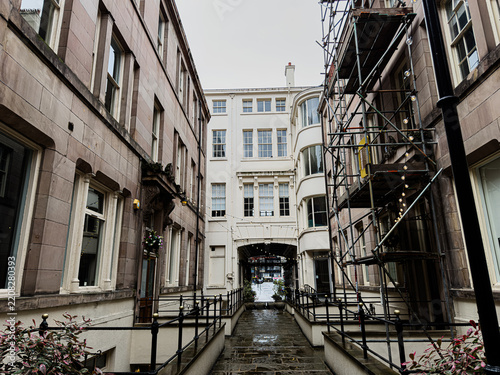 Narrow wet alleyway with historic architecture and scaffolding in Liverpool, UK, showing stone facades, iron railings and a distant street view