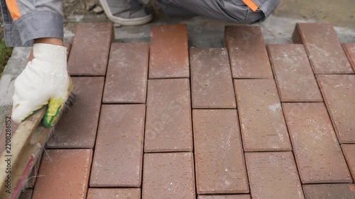 Close up video of a builder laying outdoor paving slabs. A worker in gloves uses a brush to sweep sand into the joints of red brick pavers to finish a new patio or walkway
