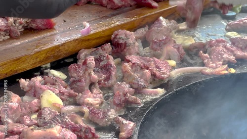 Sizzling fresh meat pieces and sliced onions grilling on a round metal plancha barbecue in bright outdoor sunlight, delicious street food culinary cooking process. 4K video.