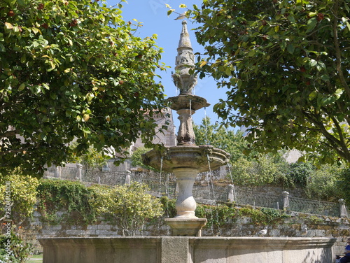 View of the historic stone fountain in Fonte Plaza de Orense, framed by lush green trees and stone walls in the city center, Pontevedra, Pontevedra, Spain.