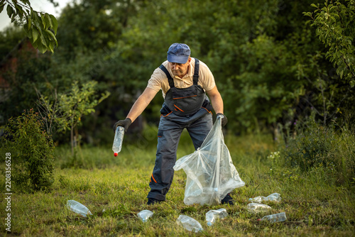 Worker picks up plastic bottle beside open trash bag in grassy, tree lined area. Focused man gathers scattered litter, supporting environmental cleanup and responsible waste disposal.