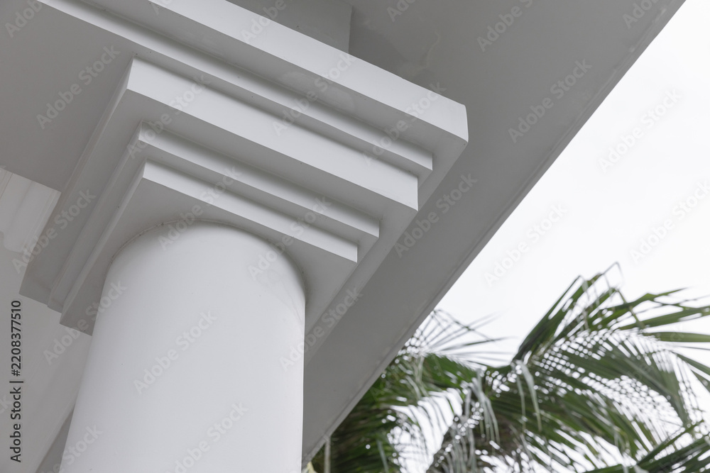 Obraz premium A white classical column with crisp molding and clean overhang on a tropical porch, framed by palm leaves. The architectural detail evokes coastal elegance, calm summer vibes
