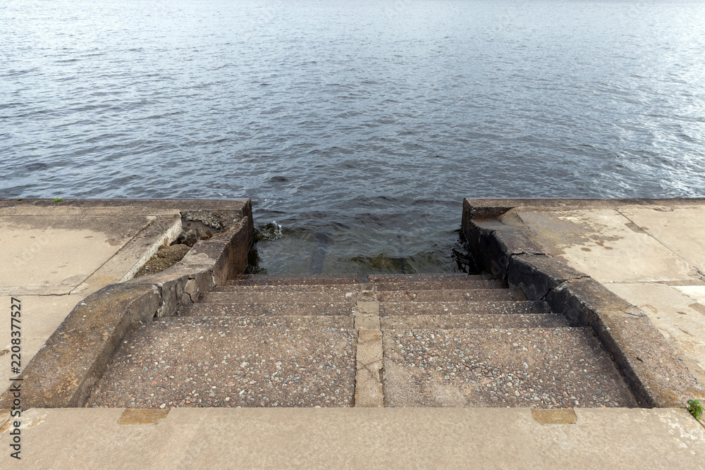 Fototapeta premium Worn concrete steps descend into calm rippling water at a weathered waterfront pier, highlighting texture, erosion and quiet atmosphere. Ideal for coastal, urban decay, travel or landscape concepts