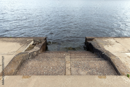 Worn concrete steps descend into calm rippling water at a weathered waterfront pier, highlighting texture, erosion and quiet atmosphere. Ideal for coastal, urban decay, travel or landscape concepts